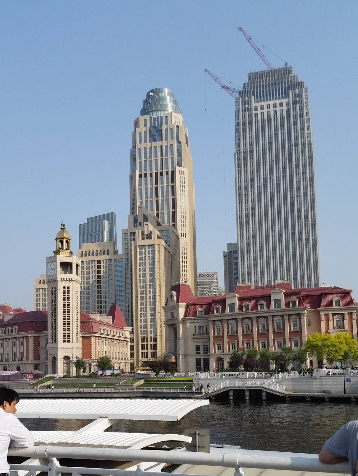 A scenic outdoor photograph of a waterfront promenade with modern high-rise buildings, a white railing in the foreground, and a clear blue sky. A person is partially visible at the bottom right, and another person is standing near the top right.